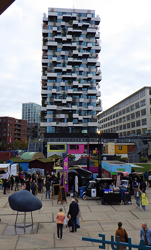 Hoge toren in Eindhoven. Nieuwbouwtoren met veel balkonnen, waar groen op geplaatst zal worden. Voor de toren staan mensen op een plein ernaar te kijken en is er een klein festival gaande.