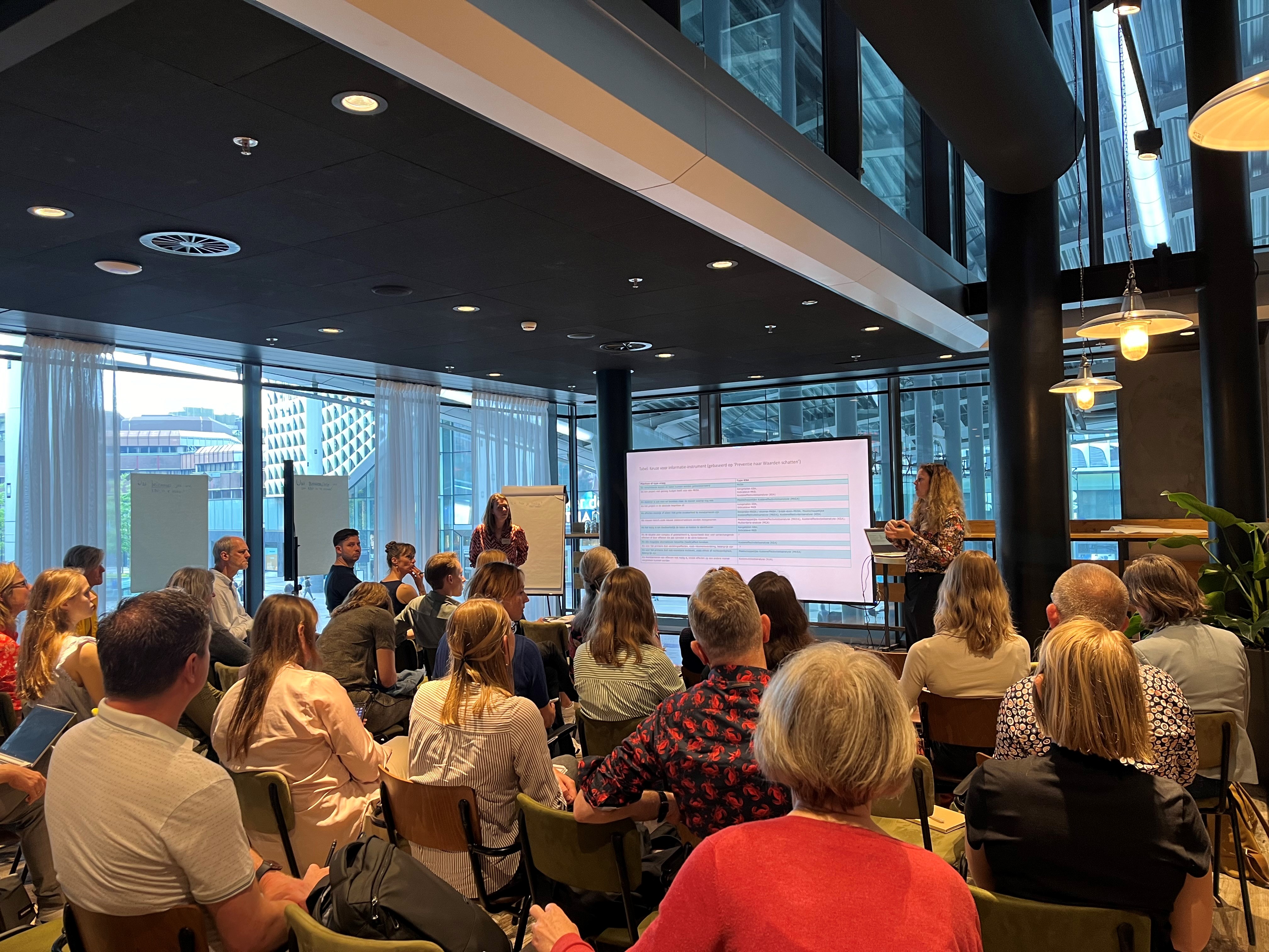 twee spreker met een presentatie zijn te zien aan de voorkant van de zaal, het publiek zit op een stoel en kijkt naar de sprekers.