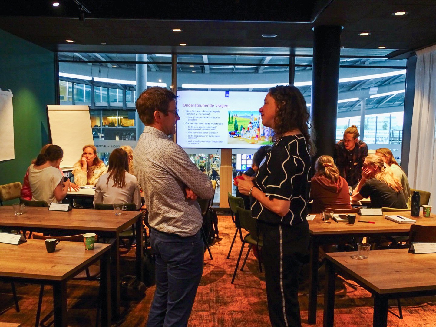 twee onderzoekers praten met elkaar voor een presentatie, op de achtergrond werken deelnemers van de workshops in groepjes aan een opdracht. 