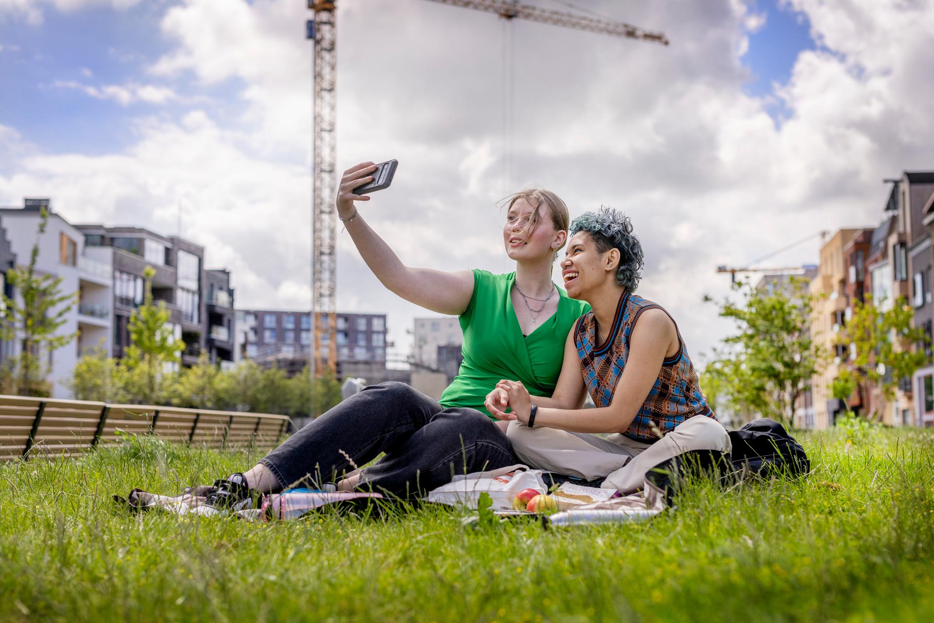 Twee vrouwen zitten naast elkaar op een kleedje op het gras en maken samen een selfie.