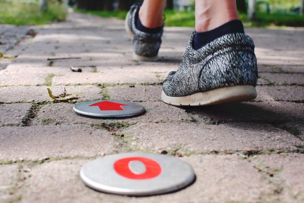 Een ingezoomde foto van twee voeten van achteren die aan het wandelen zijn. De tegels en twee afstandsindicatoren van een wandelroute zijn zichtbaar.