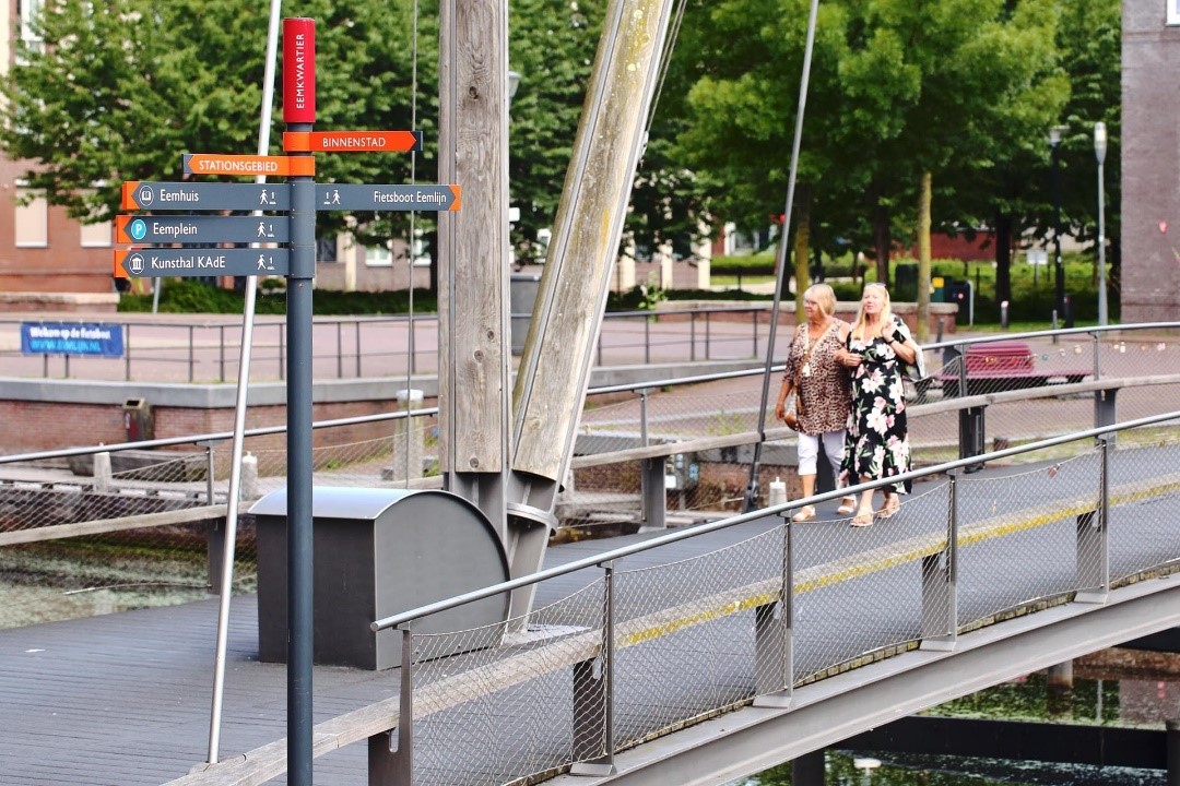 Twee dames lopen samen op een brug in Amersfoort, ze lopen richting de camera en steken de brug over. Ook is er een paal met bordjes met de trekpleisters van Amersfoort te zien.