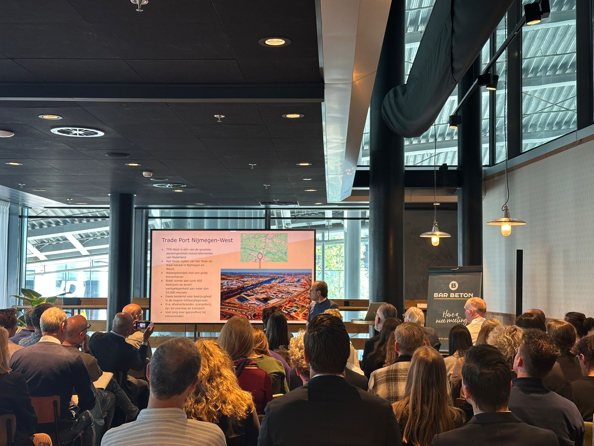 Op de afbeelding is te zien een zaal met deelnemers en de presentatie over Trade Port Nijmegen-West