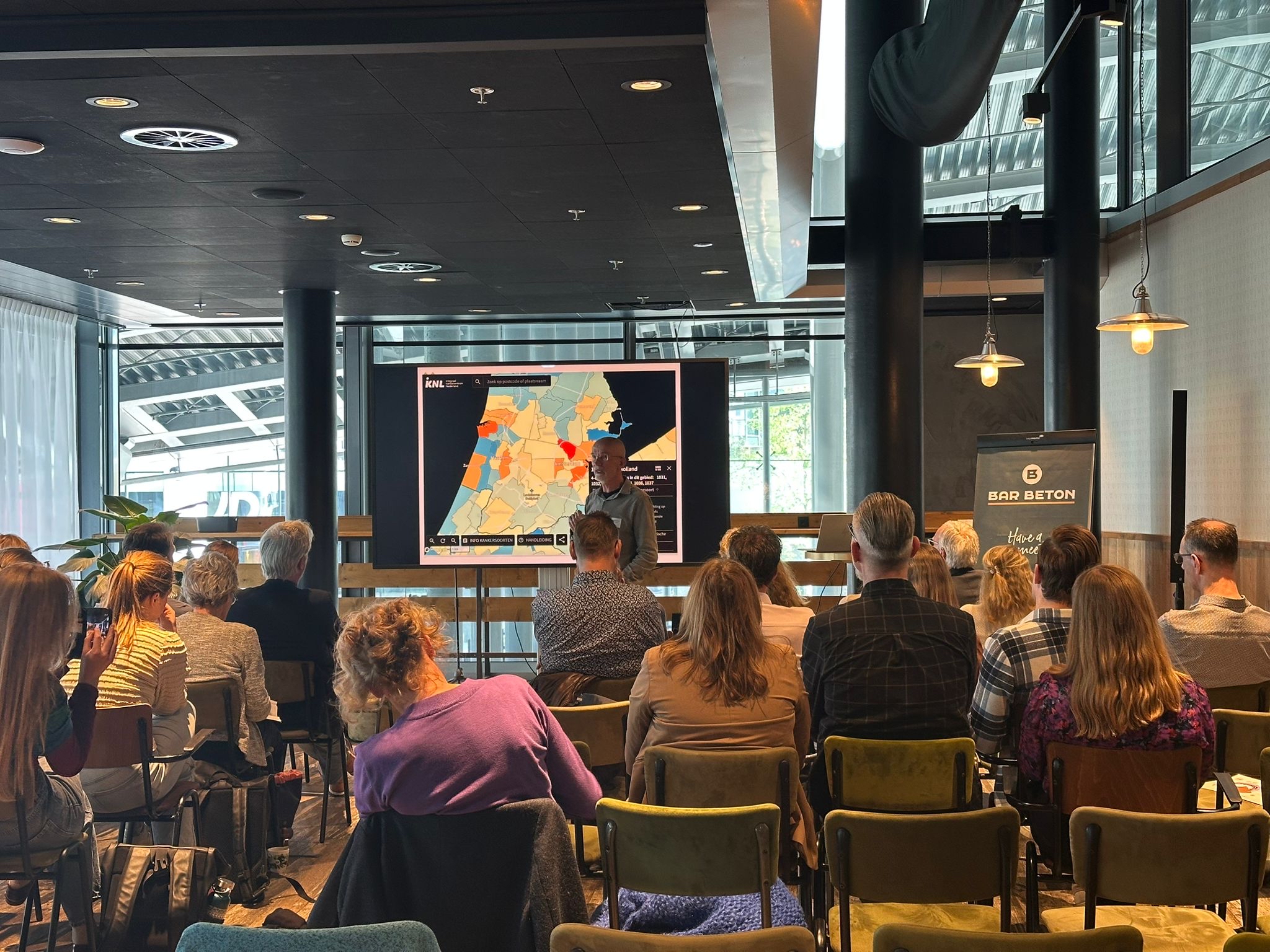 Op de afbeelding zie je de zaal met deelnemers en een presentatie