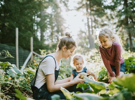 Een moeder zit met 2 kinderen gehurkt tussen groene gewassen. Ze laat iets zien wat ze aan het oogsten zijn.