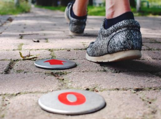 Een ingezoomde foto van twee voeten van achteren die aan het wandelen zijn. De tegels en twee afstandsindicatoren van een wandelroute zijn zichtbaar.