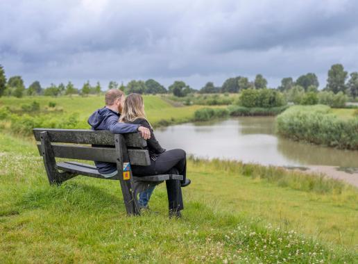 Man en vrouw zitten met hun rug naar de camera toe op een bankje kijkend over het groen en water. De omgeving is zeer groen (velden) en water is ook aanwezig.