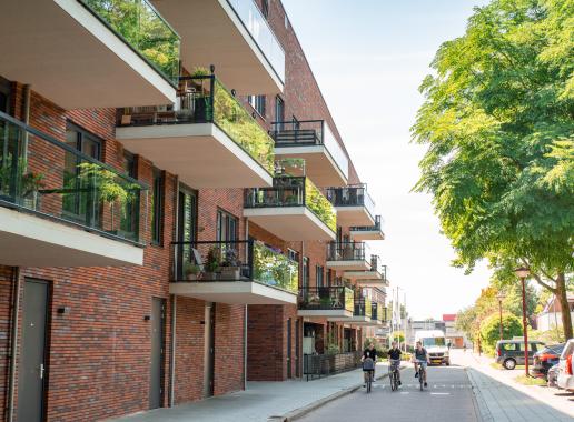 Een foto van een nieuwbouwcomplex, waaronder op straat drie kinderen de richting van de camera op komen fietsen.