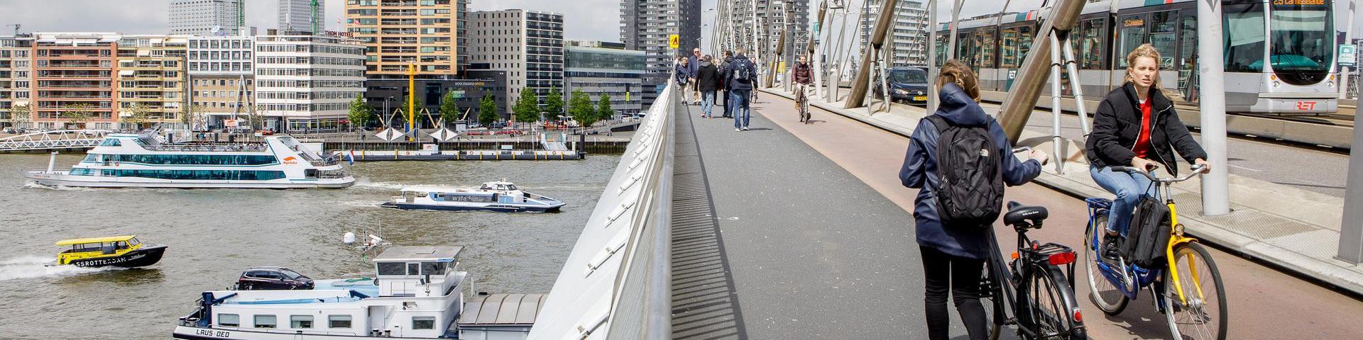 Voetgangers, fietsers en een tram op de Erasmusbrug. Onder de brug varen enkele schepen. Een mevrouw fietst op een OV-fiets.