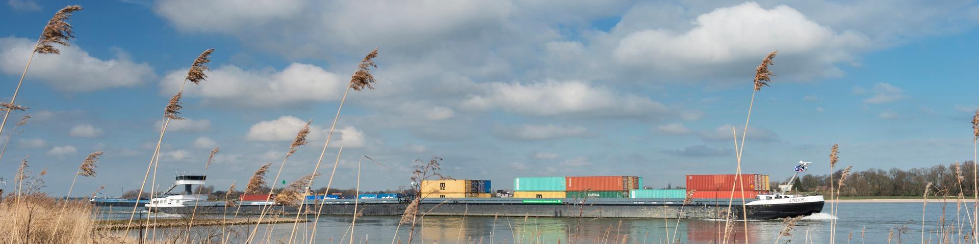 Nederland, Noord-Brabant, Gameren, maart 2014 Een binnenvaartschip met containers vaart stroomopwaarts op de rivier de Waal, riet op de voorgrond op de oever, wolkjes in de lucht.