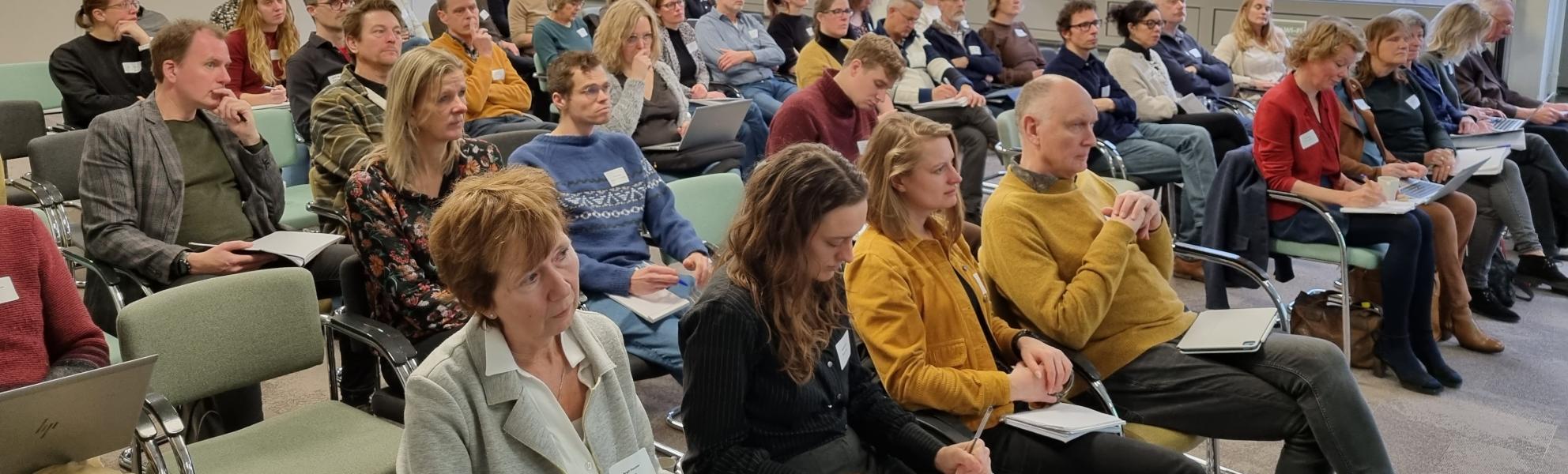 Ongeveer 50 mensen zitten in de zaal bij bijeenkomst Maak Ruimte voor Gezondheid, met hun gezicht naar ons toe, luisterend naar de spreker.