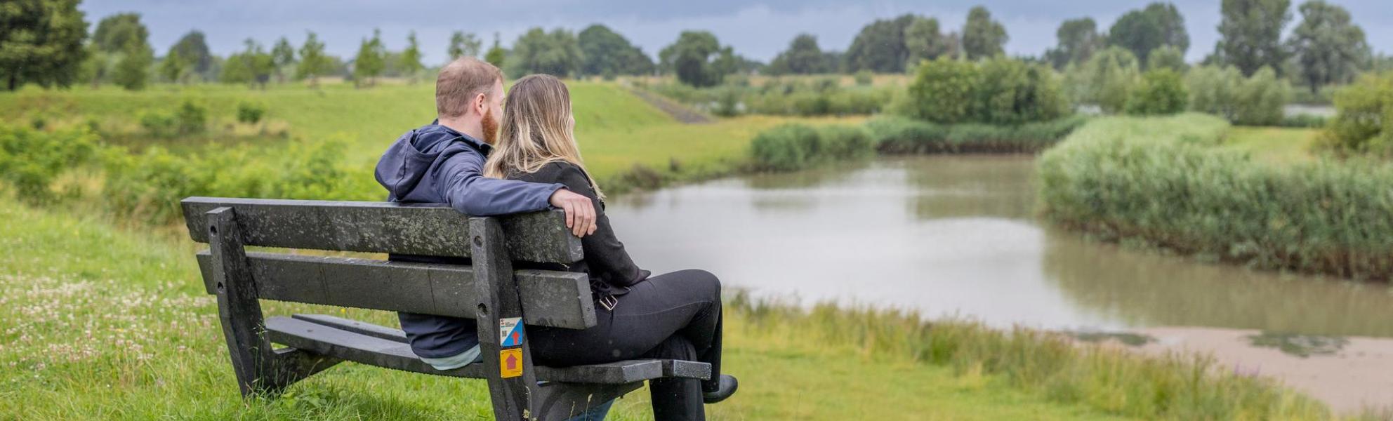 Man en vrouw zitten met hun rug naar de camera toe op een bankje kijkend over het groen en water. De omgeving is zeer groen (velden) en water is ook aanwezig.