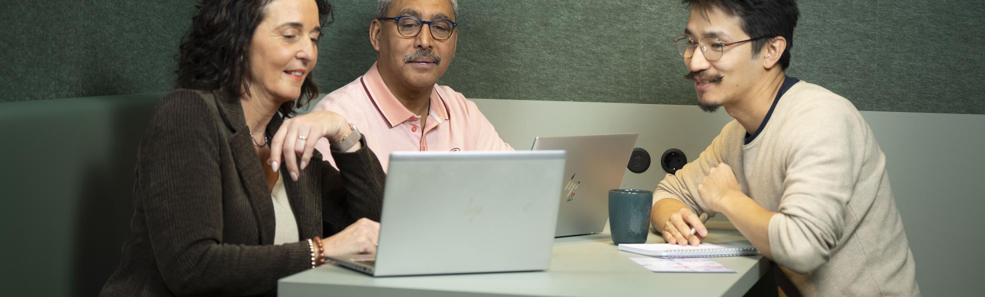 Drie personen zitten aan een tafel achter twee laptops. Ze zijn met elkaar in gesprek over wat er op het beeld staat