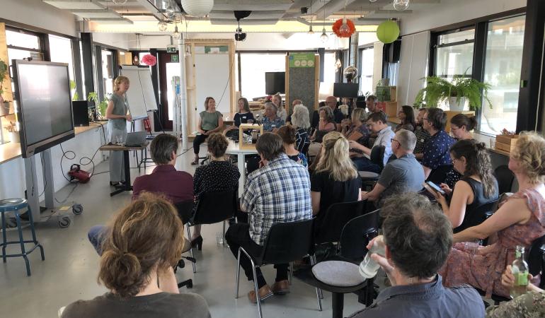 Zaal met ongeveer 40 man zittend op stoelen kijkt naar Brigit Staatsen die aan het woord is.