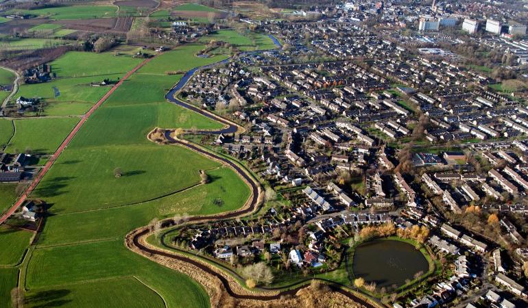 Een luchtfoto van De Dommel, een beek stroomafwaarts een kleinere rivier in de Kempen en de Meierij van 's-Hertogenbosch. Je ziet een stuk platteland en stad.