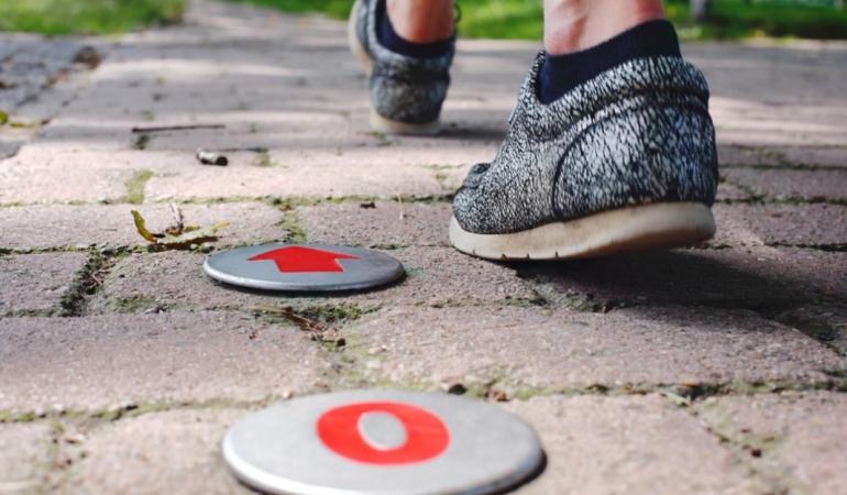 Een ingezoomde foto van twee voeten van achteren die aan het wandelen zijn. De tegels en twee afstandsindicatoren van een wandelroute zijn zichtbaar.