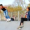 Twee licht getinte jongeren (man, vrouw) staan en zitten op de rand van een skatebaan. De jongen links heeft skeelers aan, de vrouw rechts kijkt lachend naar de man.